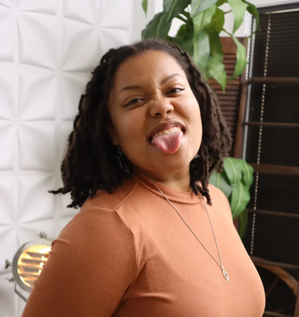 Black woman with locs in a rust-colored top playfully sticking out her tongue while standing in a cozy, plant-filled workspace—capturing the confident, creative spirit behind Olivia’s Desk, an Organic Growth Copywriter for brands that want to connect with clarity.